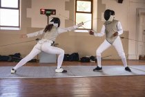 Caucasian and African American sportswomen wearing protective fencing outfits during a fencing training session, jumping taking aim at each other with their epees. Fencers training at gym. — Stock Photo