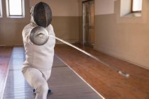 Deportista caucásico con traje de esgrima protectora durante una sesión de entrenamiento de esgrima, preparándose para un duelo, sosteniendo un pis y atacando. Esgrimistas entrenando en un gimnasio. - foto de stock