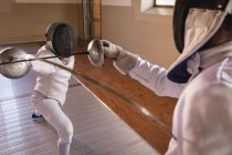 Kaukasische und afroamerikanische Sportlerinnen tragen bei einem Fechttraining Schutzanzüge und duellieren sich mit ihren Degen. Fechter trainieren im Fitnessstudio. — Stockfoto
