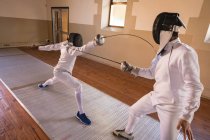 Caucasian and African American sportswomen wearing protective fencing outfits during a fencing training session, lunging at each other with their epees. Fencers training at gym. — Stock Photo