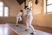 Caucasian and African American sportswomen wearing protective fencing outfit during a fencing training session, duelling with their epees. Fencers training at gym. — Stock Photo