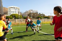 Vista lateral de dos equipos multiétnicos de jugadores de fútbol con sus tiras de equipo, en acción durante un partido de fútbol en un campo de fútbol al sol, un niño dirigiendo la pelota hacia el gol - foto de stock