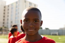 Portrait of an African American boy soccer player wearing a team strip, standing on a playing field in the sub, looking to camera and smiling, with teammates standing in the background — Stock Photo
