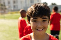 Portrait of a mixed race boy soccer player wearing his team strip, standing on a playing field in the sun, looking to camera and smiling, with teammates standing in the background — Stock Photo