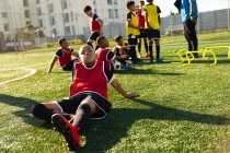Front view of a mixed race boy soccer player sitting on a playing field in the sun resting and looking to camera during a training session, with his teammates listening to their coach in the background — Stock Photo