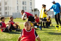 Front view of a mixed race boy soccer player sitting on a playing field in the sun resting and looking to camera during a training session, with his teammates listening to their coach in the background — Stock Photo