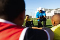 Вид спереди на футбольного тренера смешанной расы, стоящего на коленях и инструктирующего многонациональную группу мальчиков-футболистов, сидящих на игровом поле на солнце во время тренировки по футболу — стоковое фото