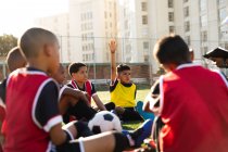 Vista lateral de un grupo multiétnico de jugadores de fútbol de niños sentados en un campo de juego en un día soleado escuchando a su entrenador masculino de raza mixta durante una sesión de entrenamiento de fútbol, un niño levantando la mano para hacer una pregunta - foto de stock