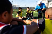 Frontansicht eines gemischten männlichen Fußballtrainers, der während eines Fußballtrainings auf einem Spielfeld in der Sonne kniet und eine multiethnische Gruppe von Jungen-Fußballern instruiert — Stockfoto