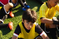 Hochwinkelporträt eines jungen Fußballspielers mit gemischter Rasse, der auf einem Spielfeld in der Sonne sitzt und während eines Fußballtrainings in die Kamera blickt, während seine Teamkollegen im Hintergrund sitzen. — Stockfoto