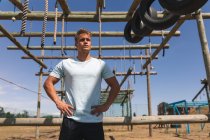 Caucasian male fitness coach at a boot camp on a sunny day, standing with hands on hips under the climbing frame of a jungle gym with hanging ropes — Stock Photo