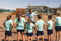 Группа кавказских мальчиков и девочек слушает инструкции кавказского тренера по фитнесу в учебном лагере в солнечный день, стоя и обращая на него внимание, пока он жестикулирует и объясняет — стоковое фото