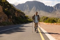Un atleta masculino de raza mixta en forma y discapacitado con pierna protésica, disfrutando de su tiempo en un viaje a las montañas, haciendo senderismo con palos, caminando por la carretera en las montañas. Estilo de vida activo con discapacidad. - foto de stock