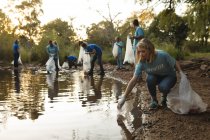 Un gruppo multietnico di volontari per la conservazione pulisce il fiume nelle campagne, raccogliendo spazzatura. Ecologia e responsabilità sociale nell'ambiente rurale. — Foto stock