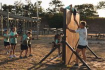 Un allenatore di fitness maschile caucasico aiuta un gruppo di ragazzi e ragazze caucasici che indossano magliette verdi e pantaloncini neri in un campo di addestramento in una giornata di sole, scavalcando una recinzione di legno in un percorso ad ostacoli — Foto stock