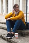 Curvy femme caucasienne dans les rues de la ville pendant la journée, assis sur des marches avec un café à emporter, parlant sur smartphone, portant un casque, avec un bâtiment historique en arrière-plan — Photo de stock