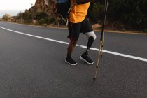 Low section of disabled male athlete with prosthetic leg, enjoying his time on a trip to the mountains, hiking with sticks, walking on the road by the sea. Active lifestyle with disability. — Stock Photo