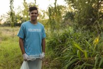 Retrato de feliz raza mixta macho voluntario de conservación limpiando el bosque en el campo, sosteniendo la bolsa de basura, sonriendo a la cámara. Ecología y responsabilidad social en el medio rural. - foto de stock
