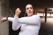 Curvy Caucasian woman with long dark hair wearing sports clothes and earphones exercising in a city, stretching her arms and warming up before her workout — Stock Photo