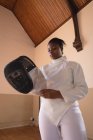 African American sportswoman wearing protective fencing outfit during a fencing training session, preparing for a duel, holding a fencing mask. Fencers training at a gym. — Stock Photo