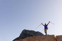 Fit, behinderter Mixed Race männlicher Athlet mit Beinprothese, genießt seine Zeit bei einem Ausflug in die Berge, beim Wandern auf den Felsen, die Arme mit Stöcken über dem Kopf. Aktiver Lebensstil mit Behinderung. — Stockfoto