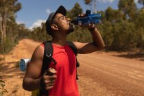 Ein fitter, behinderter Mixed-Race-Athlet mit Beinprothese, der seine Zeit beim Wandern genießt, auf einem Feldweg im Wald steht und Wasser trinkt. Aktiver Lebensstil mit Behinderung. — Stockfoto