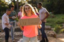 Portrait of of happy Caucasian female conservation volunteer cleaning up the countryside with board Our Future, her friends in the background. Ecology and social responsibility in rural environment. — Stock Photo