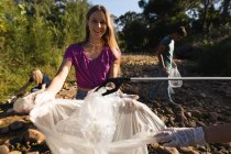 Retrato de una voluntaria de conservación caucásica limpiando el río en el campo, sus amigas recogiendo basura en el fondo. Ecología y responsabilidad social en el medio rural. - foto de stock