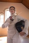 Femme sportive caucasienne portant une tenue d'escrime protectrice lors d'une séance d'entraînement d'escrime, se préparant à un duel, tenant une épee et un masque. Entraînement des escrimeurs dans un gymnase. — Photo de stock