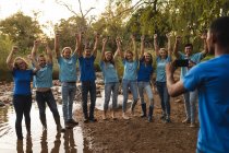 Мужчина фотографирует счастливую многонациональную группу волонтеров по охране природы, очищающих реку в сельской местности, улыбаясь с поднятыми руками. Экология и социальная ответственность в сельской местности. — стоковое фото