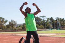 Fit, mixed race disabled male athlete at an outdoor sports stadium, kneeling on race track after race with arms in the air wearing running blades. Disability athletics sport training. — Stock Photo