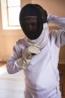 Кавказский спортсмен в защитной одежде во время тренировки по фехтованию, готовится к поединку, надевает маску. Обучение фехтовальщиков в спортзале. — стоковое фото