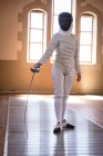 Esportista caucasiana vestindo roupa de esgrima protetora durante uma sessão de treinamento de esgrima, se preparando para um duelo, segurando um epee. Treinamento de esgrimistas em um ginásio. — Fotografia de Stock