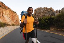 Athlète masculin de race mixte en forme et handicapé avec une jambe prothétique, profitant de son temps sur un voyage à la montagne, randonnée avec des bâtons, marche sur la route par la mer. Mode de vie actif avec handicap. — Photo de stock
