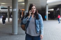 Kurvige kaukasische Frau, die tagsüber auf den Straßen der Stadt unterwegs ist, lächelt und ihr Smartphone benutzt, einen Rucksack trägt und mit einem modernen Gebäude im Hintergrund spaziert — Stockfoto