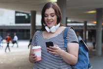 Curvy Caucasian woman out and about in the city streets during the day, smiling, using her smartphone wearing headphones and backpack, holding a takeaway coffee, with modern building in the background — Stock Photo