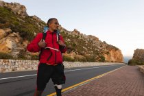 Un atleta masculino en forma, discapacitado de raza mixta con pierna protésica, disfrutando de su tiempo en un viaje a las montañas, senderismo, caminar por la carretera junto al mar. Estilo de vida activo con discapacidad. - foto de stock