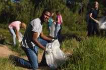 Ritratto di un volontario afroamericano di conservazione che ripulisce il fiume in campagna, mentre i suoi amici raccolgono spazzatura sullo sfondo. Ecologia e responsabilità sociale nell'ambiente rurale. — Foto stock