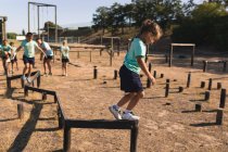 Caucasian girl at a boot camp on a sunny day, wearing green t shirt and black shorts, balancing and walking along a beam on an obstacle course, with other kids following her in the background — Stock Photo