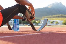 Low section of fit, disabled male athlete at an outdoor sports stadium, resting with water bottle on race track wearing running blades. Disability athletics sport training. — Stock Photo