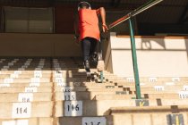 Vista trasera del atleta masculino discapacitado de carrera mixta en un estadio de deportes al aire libre, subiendo escaleras en las gradas con auriculares y cuchillas para correr. Discapacidad atletismo entrenamiento deportivo. - foto de stock