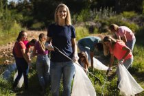 Retrato de uma voluntária caucasiana de conservação limpando o rio no campo, seus amigos pegando lixo no fundo. Ecologia e responsabilidade social no meio rural. — Fotografia de Stock