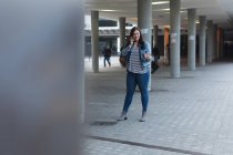 Curvy femme caucasienne dans les rues de la ville pendant la journée, souriant, gesticulant et parlant sur son smartphone, marchant avec un bâtiment moderne en arrière-plan — Photo de stock