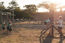 Un entrenador de fitness masculino caucásico que ayuda a un grupo de niños y niñas caucásicos que usan camisetas verdes y pantalones cortos negros en un campamento de entrenamiento en un día soleado, trepando por una valla de madera en una carrera de obstáculos - foto de stock
