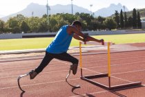 Fit, mixed race disabled male athlete at an outdoor sports stadium, preparing before workout stretching on hurdle on race track wearing running blades. Disability athletics sport training. — Stock Photo