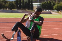 Fit, mixed race disabled male athlete at an outdoor sports stadium, sitting on race track after race checking his smartwatch with water bottle wearing running blades. Disability athletics sport — Stock Photo