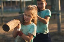Кавказская девушка и мальчик в грязно-зеленых футболках и черных шортах, несущих бревно вместе на своих плечах во время тренировки в учебном лагере в солнечный день — стоковое фото