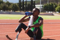 Fit, gemischt rennbehinderter männlicher Athlet in einem Outdoor-Sportstadion, sitzt nach dem Rennen auf der Rennstrecke und trinkt Wasser mit laufenden Klingen. Behindertensport. — Stockfoto