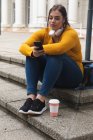 Mujer curvilínea caucásica por las calles de la ciudad durante el día, sentada en escalones con un café para llevar, usando su teléfono inteligente, usando auriculares, con un edificio histórico en el fondo - foto de stock