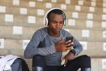 Fit, mixed race disabled male athlete at an outdoor sports stadium, sitting in the stands wearing headphones using smartphone wearing running blades. Disability athletics sport training. — Stock Photo